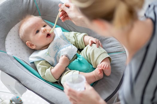 Mother spoon feeding her baby boy child in baby chair with fruit puree. Baby solid food introduction concept