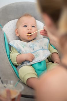 Mother spoon feeding her baby boy child in baby chair with fruit puree. Baby solid food introduction concept