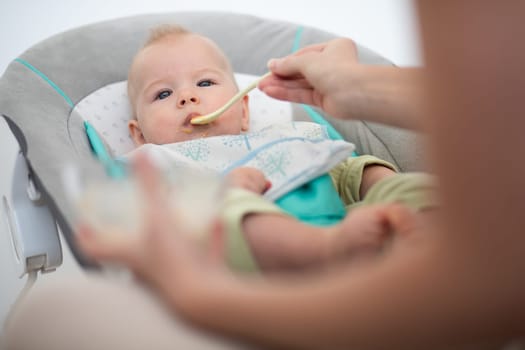 Mother spoon feeding her baby boy child in baby chair with fruit puree. Baby solid food introduction concept