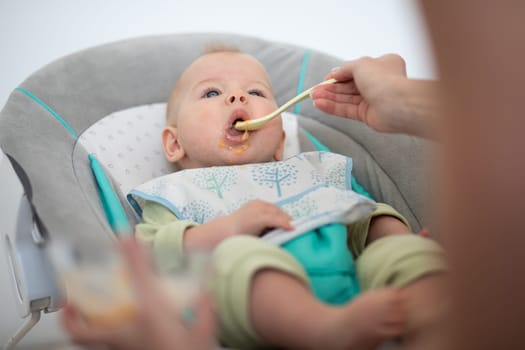 Mother spoon feeding her baby boy child in baby chair with fruit puree. Baby solid food introduction concept