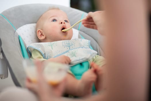 Mother spoon feeding her baby boy child in baby chair with fruit puree. Baby solid food introduction concept