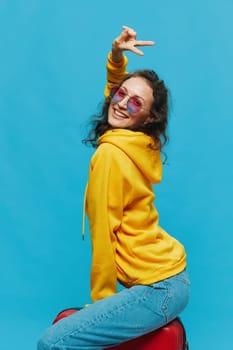 Smile woman sitting on a suitcase in a yellow hoodie, blue jeans and glasses on a blue background, packing for a trip. High quality photo