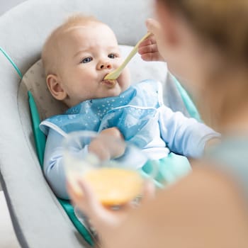 Mother spoon feeding her baby boy child in baby chair with fruit puree. Baby solid food introduction concept
