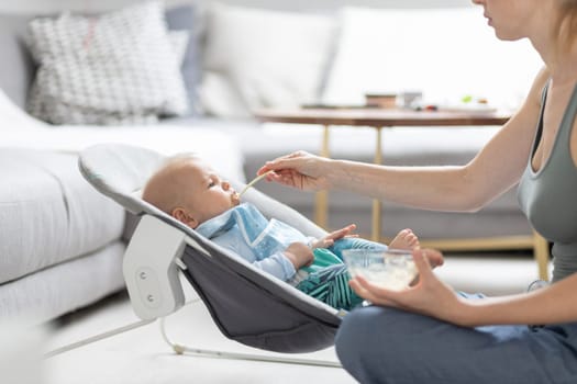 Mother spoon feeding her baby boy child in baby chair with fruit puree. Baby solid food introduction concept