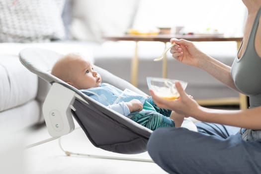 Mother spoon feeding her baby boy child in baby chair with fruit puree. Baby solid food introduction concept