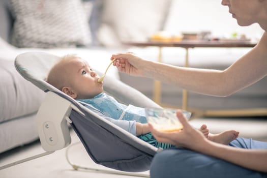 Mother spoon feeding her baby boy child in baby chair with fruit puree. Baby solid food introduction concept