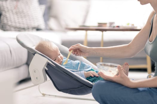 Mother spoon feeding her baby boy child in baby chair with fruit puree. Baby solid food introduction concept