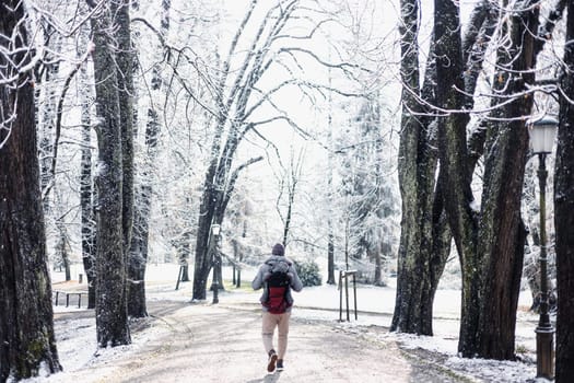 Rear view of sporty father carrying his infant son wearing winter jumpsuit and cap in backpack carrier walking in city park in winter.