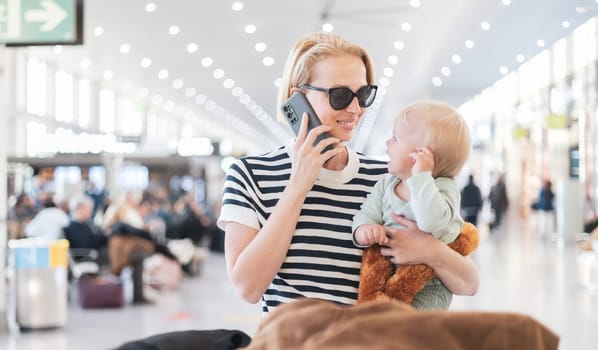 Mother talking on mobile phone while traveling with child, holding his infant baby boy at airport terminal waiting to board a plane. Travel with kids concept