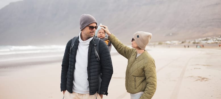 Young father carrying his infant baby boy son in backpack on windy sandy Famara beach, Lanzarote island, Spain. Family travel and winter vacation concept