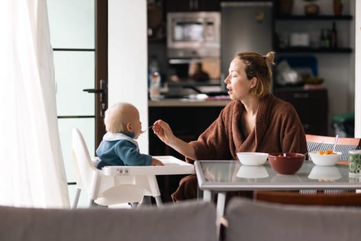 Mother wearing bathrope spoon feeding her infant baby boy child sitting in high chair at the dining table in kitchen at home in the morning.