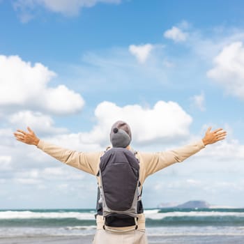 Young father rising hands to the sky while enjoying pure nature carrying his infant baby boy son in backpack on windy sandy beach of Famara, Lanzarote island, Spain. Family travel concept