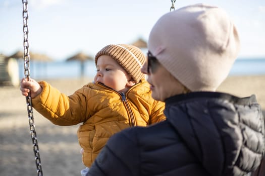 Mother pushing her infant baby boy child on a swing on sandy beach playground outdoors on nice sunny cold winter day in Malaga, Spain