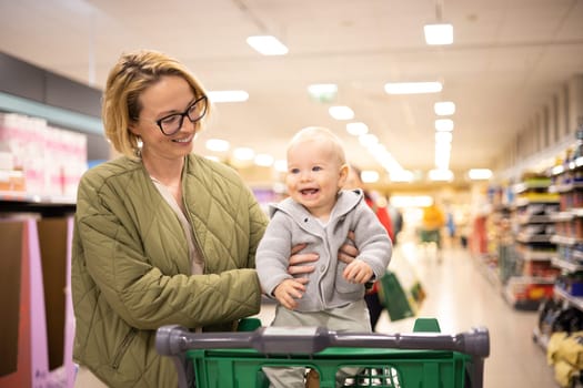 Mother pushing shopping cart with her infant baby boy child down department aisle in supermarket grocery store. Shopping with kids concept