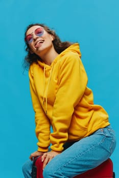 Smile woman sitting on a suitcase in a yellow hoodie, blue jeans and glasses on a blue background, packing for a trip. High quality photo