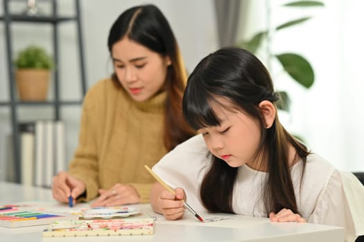 Adorable little schoolgirl doing homework, drawing with happy mother at home.