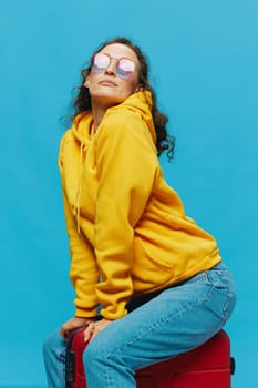 Smile woman sitting on a suitcase in a yellow hoodie, blue jeans and glasses on a blue background, packing for a trip. High quality photo