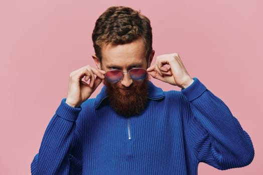 Portrait of a redheaded man wearing sunglasses smiling and dancing, listening to music on a pink background. Hipster with a beard, happiness finger pointing. High quality photo