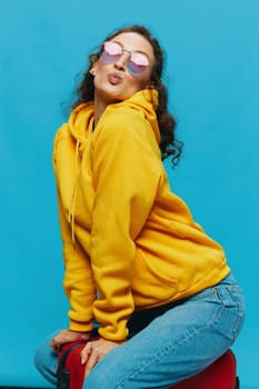 Smile woman sitting on a suitcase in a yellow hoodie, blue jeans and glasses on a blue background, packing for a trip. High quality photo