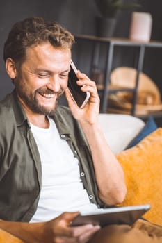 Happy man enjoying phone call while holding smartphone in one hand and tablet PC in other. He seat comfortably home, taking advantage of modern technology and convenience of being able to multitask. High quality photo