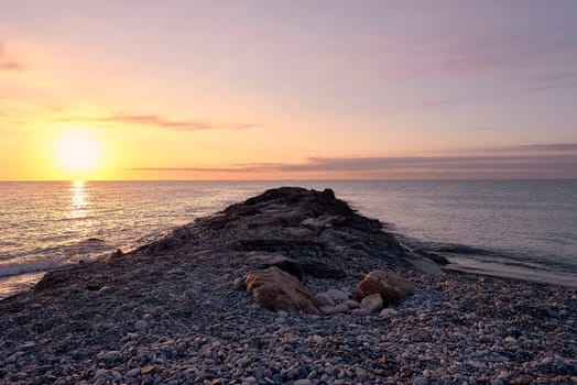 Breakwater on the rocky beach at sunrise. Lonely beach, sunshine, orange sky, small stones, calm sea