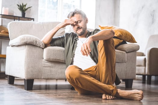 Middle-aged man resting on floor next to sofa home, moment of contemplation or crisis. Difficulties and challenges faced by men in society as they age, with focus on theme of middle age and its associated struggles. High quality photo