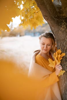 The blonde stands near the autumn tree. Thoughtful woman looks ahead, dressed in a yellow dress. Autumn content.