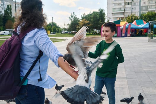 Lifestyle portrait of a Caucasian happy teen boy in casual clothes, smiling cheerfully and having fun while walking with his mother and feeding pigeons in the park. People. Carefree childhood. Animals