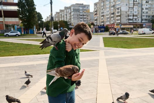 Smiling adorable Caucasian teen boy, expressing positive emotions while feeding rock pigeons and feral doves in the urban square. The concept of people, nature and kindness for animals