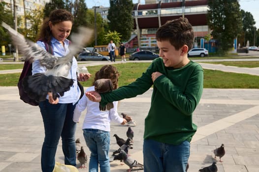 Happy multi ethnic family of loving mother and two kids feeding pigeons in the square of a city park. People. Nature. Human relationships. Family leisures and outing. The concept of care for animals