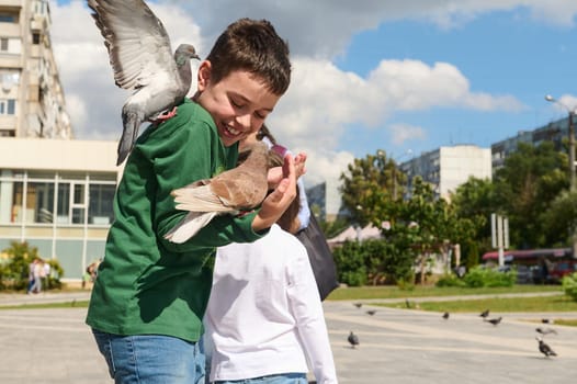 Caucasian child boy expressing happy positive emotions while walking on the city square and feeding pigeons flying to his back. People. Happy and carefree childhood. Animals and nature concept