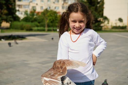 Authentic portrait of adorable Caucasian little girl 5 years old with two ponytails, smiling with a beautiful cheerful toothy smile looking at camera, standing in the square and feeding rock pigeons