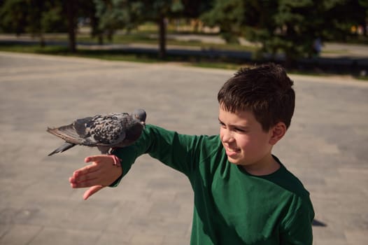 Authentic portrait of adorable multi ethnic pre teen boy, smiling and having fun while standing in the square and feeding rock pigeons. Carefree and happy hildhood. People and wild animals concept