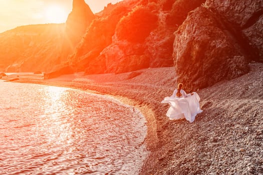 Mysterious woman silhouette long hair walks on the beach ocean water, sea nymph wind listens to the wave. Throws up a long white dress, a divine sunset. Artistic photo from the back without a face.