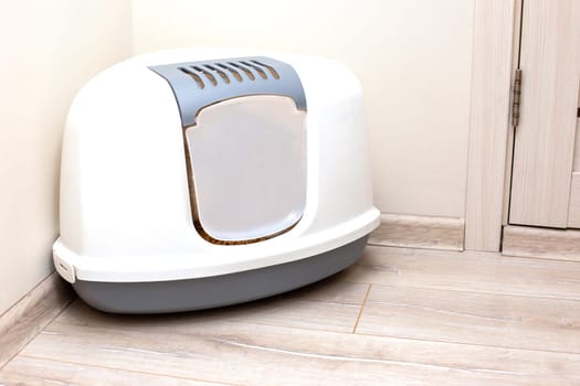 A large, gray, triangular plastic, enclosed litter box for a cat sits on the floor in the corner of a bright room.