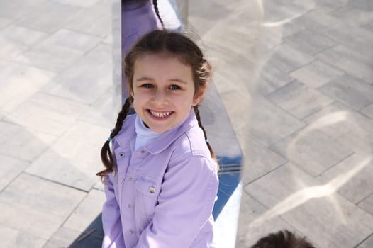 Authentic portrait of Caucasian happy delightful child girl 5 years old, smiling with beautiful cheeerful toothy smile, looking at camera, against gray urban background. View from above. Copy space