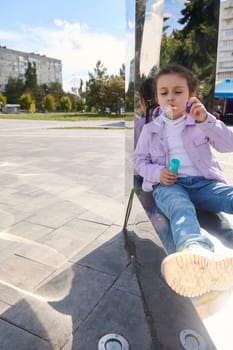 Caucasian adorable child 5 years old, charming little girl in casual denim, sitting on mirror metal bench and blowing soap bubbles in city park. Childhood. Family outing. Leisure activity. Urban life