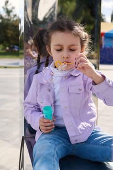 Caucasian little kid girl 5-6 years old, wearing purple jacket and blue jeans, blowing soap bubbles while resting in city park, sitting on a metal mirror bench. Leisure activity. Lifestyle. Urban life