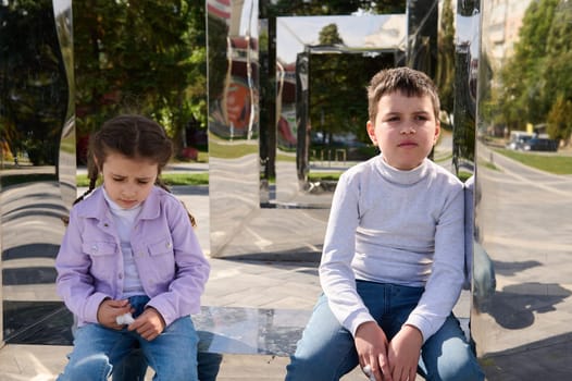 Portrait of two adorable elementary aged kids, boy and girl, brother and sister sitting on mirror bench in urban park. Family outing. Friendships and human relationships. People. Lifestyle. Leisures