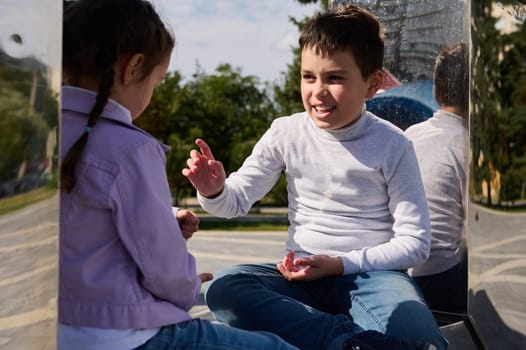 Lovely pre teen boy talking with his little sister while relaxing sitting on mirror bench on a sunny warm spring day. Brother and sister enjoying a happy weekend together in urban park. Family outing