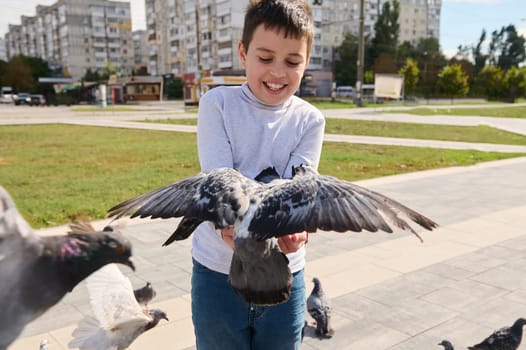 Adorable Caucasian child boy 10 years old, having fun during family outing, feeding flock of flying doves to his hands, taking care for animals in the town square. Childhood, wild animals and nature
