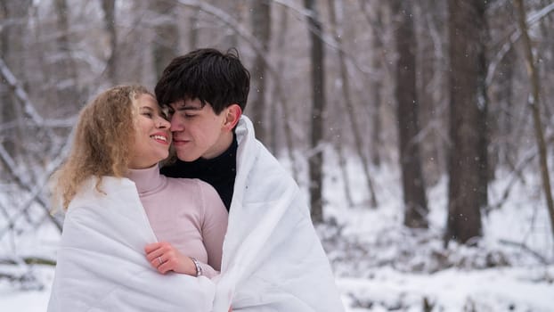 A young couple walks in the park in winter. The guy and the girl are kissing wrapped in a white blanket outdoors