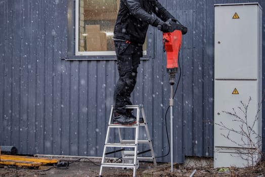 A worker installs a ground rod to ground a building. A worker in work clothes drives earth rods into the ground with a jackhammer to prevent short circuits