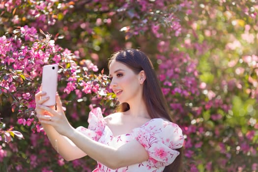 A beautiful brunette girl with long hair is standing near a pink blooming apple tree, in the summer in the park, on a sunny day, holding a pink smartphone, taking pictures of herself. Close up