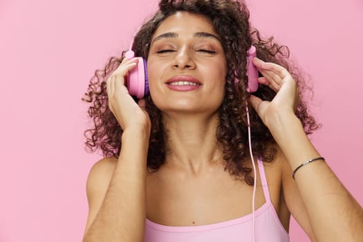 Happy woman wearing headphones with curly hair listening to music and singing along with her eyes closed in a pink T-shirt and jeans on a pink background, copy space. High quality photo