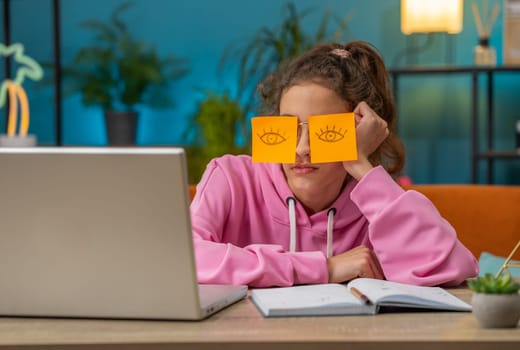 Tired Caucasian preteen school girl sleeping on laptop computer with eyes stickers on face. Young lazy child, kid cheating to sleep at home workplace sitting at the table. Distance learning education