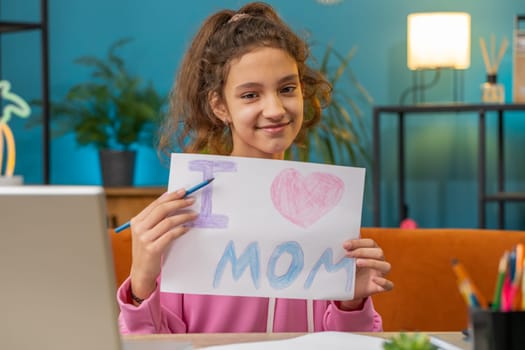 Preteen school girl drawing colorful picture I Love Mom with pencils on white paper, greeting card surprise for mother. Happy Mother's Day. Young child, kid at home workplace sitting at the table
