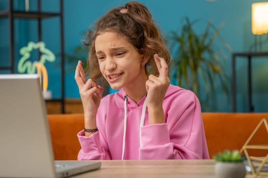 Caucasian preteen school girl looking at laptop screen crossed fingers wishing good exam results, dreaming about win victory in lottery jackpot game. Child kid at home workplace sitting at the table