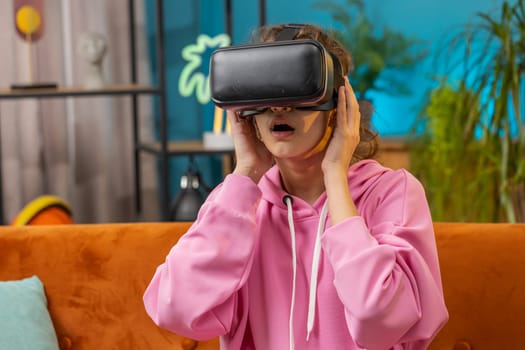 Preteen school girl using virtual reality futuristic technology VR app headset helmet to play simulation 3D 360 video game, watching film movie. Child, kid at home apartment sitting on orange couch