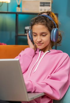 Caucasian preteen school girl in headphones use laptop listens to music or lesson, distance learning. Online education. Child kid relaxing, taking a break at home workplace sitting on couch. Vertical